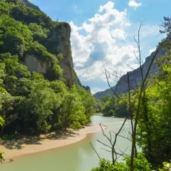 La **Gola del Furlo** vista dal fiume, con pareti rocciose ricoperte di vegetazione e un cielo azzurro con nuvole bianche.