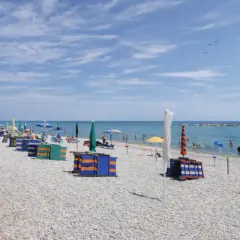 Spiaggia di ciottoli con ombrelloni colorati e cabine, bagnanti in mare. Un tranquillo fermo mare sotto un cielo azzurro con nuvole leggere.