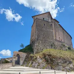 Antico borgo di Frontone, Marche, con imponente rocca in pietra su un colle erboso, sotto un cielo azzurro con nuvole bianche.