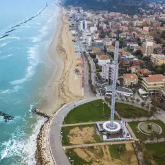 Vista aerea del lungomare di Pesaro con la spiaggia, il mare con frangiflutti e una torre panoramica.