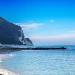 Scogliera a picco sul mare azzurro, spiaggia dorata e gente che passeggia sulla riviera marchigiana.