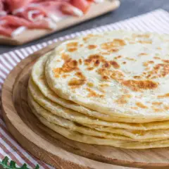 Una pila di cresce dorate su un tagliere di legno, pronte per essere farcite con rucola fresca e prosciutto.