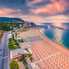 Vista aerea della spiaggia numana con file ordinate di ombrelloni rossi e blu, mare calmo e cielo rosa al tramonto.