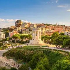 Panoramica di un **paesaggio** collinare nelle **Marche**, con un monumento bianco al centro, case colorate sullo sfondo e un parco verdeggiante.