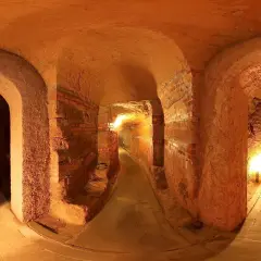 Un labirinto di corridoi illuminati, con archi in mattoni e terra battuta, che si estende in profondità. Sembra di esplorare antiche grotte sotterranee.