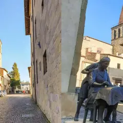 Sansepolcro cosa vedere: una piazza con una scultura di donne al lavoro, campanili medievali e palazzi storici sotto un cielo azzurro.
