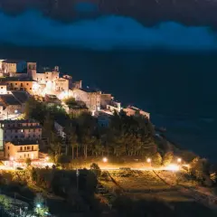 Norcia: cosa vedere dopo il terremoto. Borgo illuminato di notte, arroccato su un colle, con case in pietra e alberi.