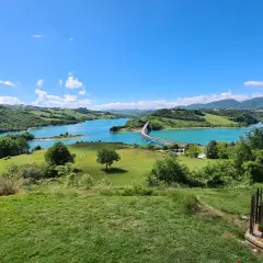 Panoramica della diga di Cingoli con lago turchese, colline verdi e un ponte ad arco sotto un cielo azzurro.