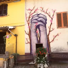Braccano, paese dei murales: mani giunte e fiori di ciliegio dipinti su un muro, con panchine e decorazioni in legno.