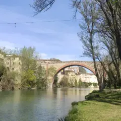 Il fiume scorre placido sotto il ponte ad arco del canyon di Fossombrone, con case antiche aggrappate alla collina e alberi che incorniciano la scena.