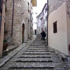 Una donna sale una scalinata in pietra in un vicolo di Gualdo Tadino, tra antiche case in pietra e intonaco.