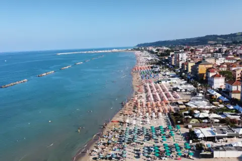 Spiaggia affollata a Porto San Giorgio, con file di ombrelloni colorati, bagnanti in mare e la città sullo sfondo.