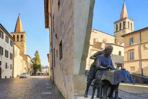 Sansepolcro cosa vedere: una piazza con una scultura di donne al lavoro, campanili medievali e palazzi storici sotto un cielo azzurro.