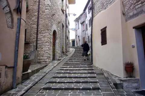 Una donna sale una scalinata in pietra in un vicolo di Gualdo Tadino, tra antiche case in pietra e intonaco.