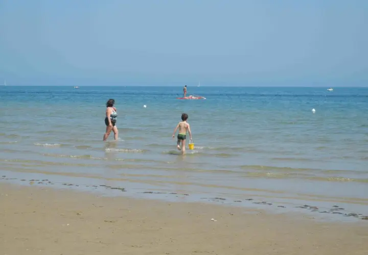 Bambino con secchiello giallo corre verso il mare a Pesaro, mentre una donna cammina nell'acqua. In lontananza, un uomo su una tavola da SUP.