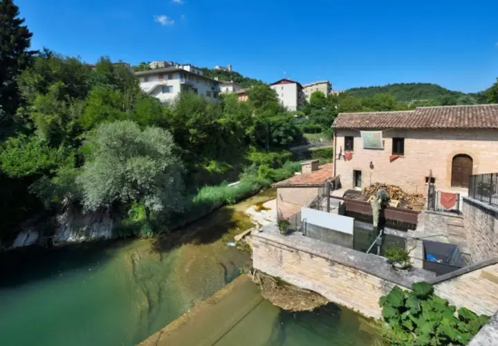 Sassoferrato cosa vedere: un antico mulino ad acqua lungo un fiume, con case e vegetazione lussureggiante sullo sfondo.