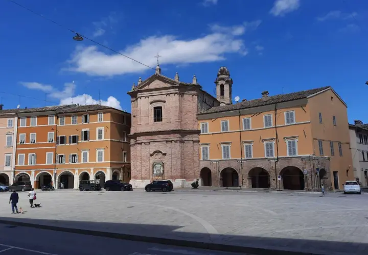 Piazza a San Severino Marche con edifici colorati, una chiesa in mattoni e cielo azzurro.