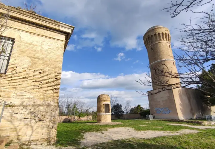 Torri antiche e un piccolo edificio circolare nel parco del cardeto, sotto un cielo azzurro con nuvole bianche.