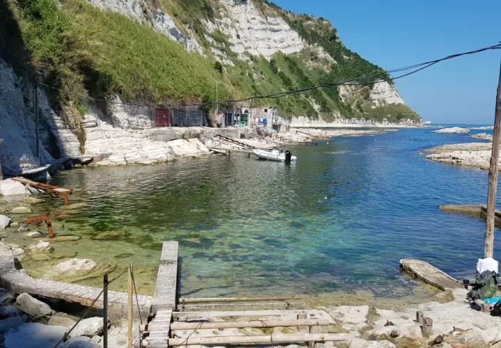 Scogli bianchi e mare cristallino alla spiaggia della Scalaccia. Una piccola barca è ormeggiata vicino a casotti colorati.