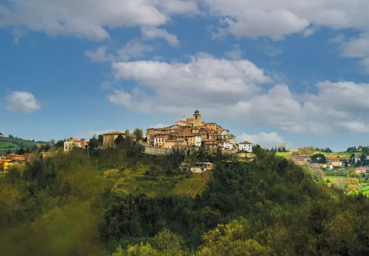 Borgo antico su un colle, con case in pietra e tetti rossi, sotto un cielo azzurro con nuvole bianche. Il **monte Grimano** domina il paesaggio.