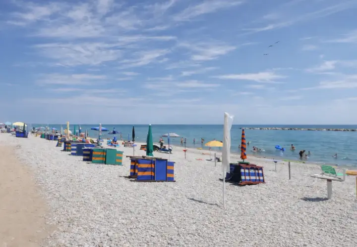 Spiaggia di ciottoli con ombrelloni colorati e cabine, bagnanti in mare. Un tranquillo fermo mare sotto un cielo azzurro con nuvole leggere.