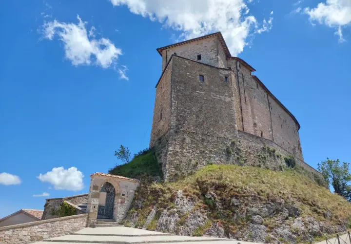 Antico borgo di Frontone, Marche, con imponente rocca in pietra su un colle erboso, sotto un cielo azzurro con nuvole bianche.
