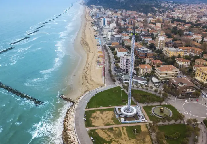 Vista aerea del lungomare di Pesaro con la spiaggia, il mare con frangiflutti e una torre panoramica.