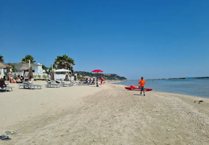 Sole splendente sulla campofilone spiaggia, con bagnanti rilassati, ombrelloni e un bagnino pronto a intervenire vicino a una barca di salvataggio.