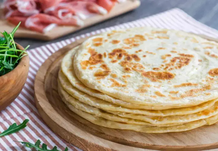 Una pila di cresce dorate su un tagliere di legno, pronte per essere farcite con rucola fresca e prosciutto.