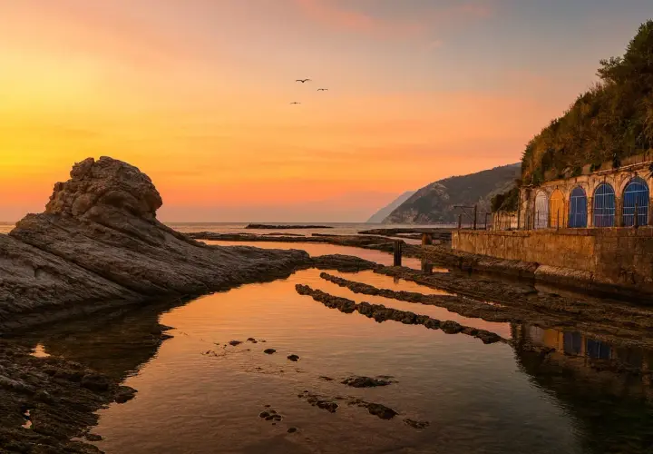 Tramonto dorato sulla spiaggia del Passetto, con rocce scolpite e archi colorati che si riflettono nell'acqua calma.