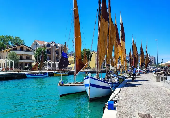Barche a vela ormeggiate in un porto con case colorate sullo sfondo. Un dilemma: meglio Bellaria o Igea Marina?