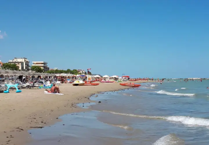 Spiaggia affollata a Cupra Marittima, con ombrelloni, lettini, bagnanti e barche di salvataggio. Il mare azzurro invita a un tuffo.