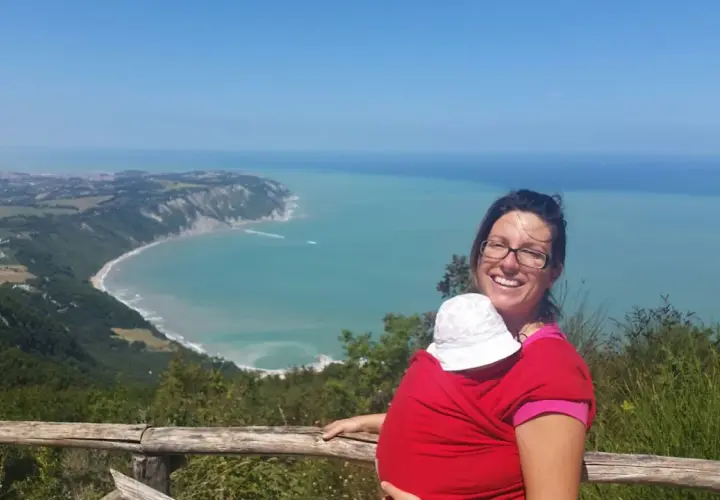 Donna sorridente con bimbo in fascia, ammira la costa del Conero dai sentieri. Perfetto per esplorare i sentieri del Conero con bambini.