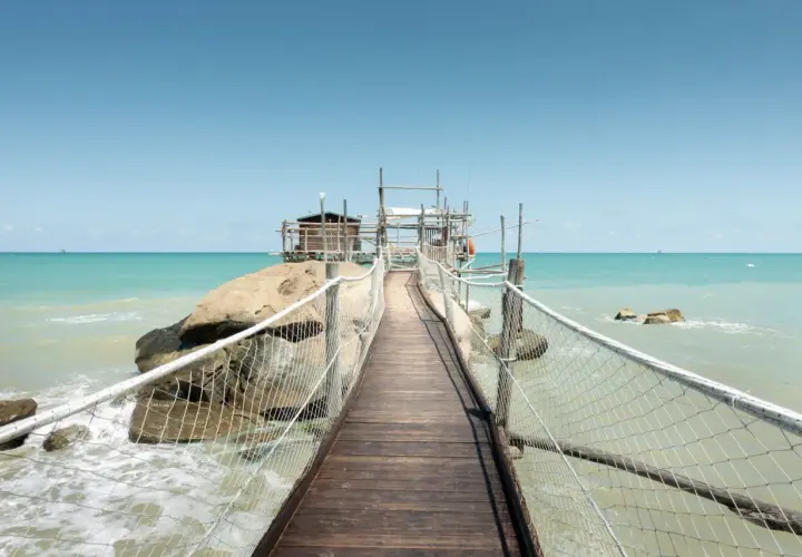 Ponte in legno verso un trabucco, tipica struttura da pesca delle Marche, su un mare azzurro e limpido.
