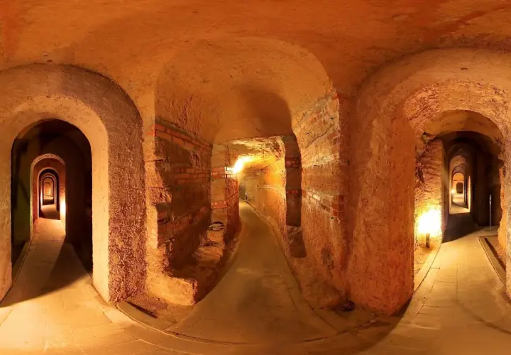 Un labirinto di corridoi illuminati, con archi in mattoni e terra battuta, che si estende in profondità. Sembra di esplorare antiche grotte sotterranee.