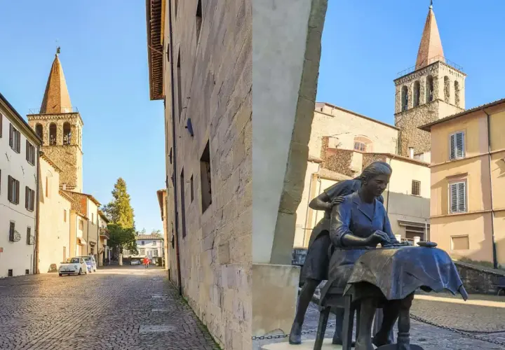 Sansepolcro cosa vedere: una piazza con una scultura di donne al lavoro, campanili medievali e palazzi storici sotto un cielo azzurro.