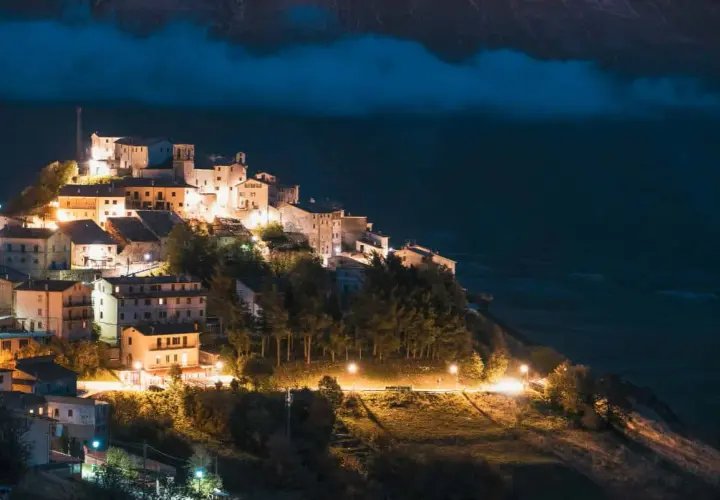 Norcia: cosa vedere dopo il terremoto. Borgo illuminato di notte, arroccato su un colle, con case in pietra e alberi.