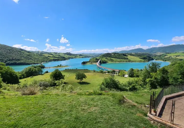 Panoramica della diga di Cingoli con lago turchese, colline verdi e un ponte ad arco sotto un cielo azzurro.