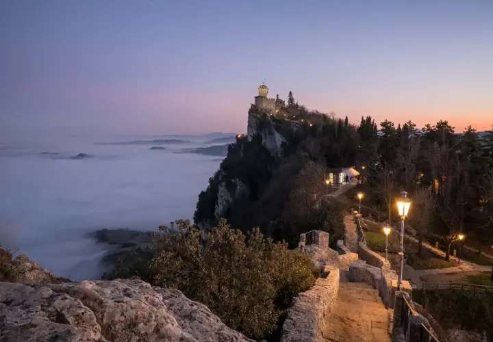 Torre Guaita domina la nebbia al tramonto, un invito a visitare San Marino per un'esperienza magica.