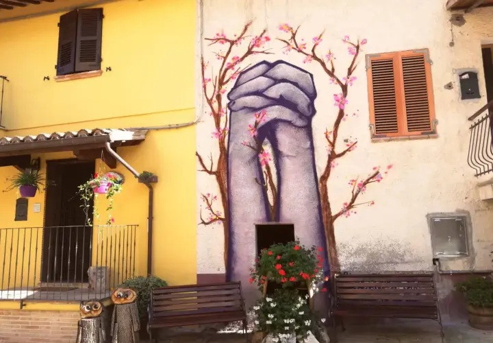 Braccano, paese dei murales: mani giunte e fiori di ciliegio dipinti su un muro, con panchine e decorazioni in legno.