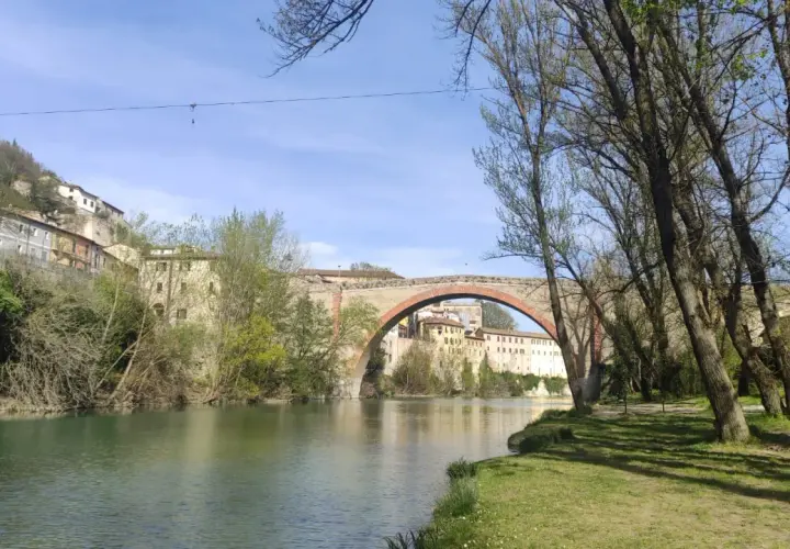 Il fiume scorre placido sotto il ponte ad arco del canyon di Fossombrone, con case antiche aggrappate alla collina e alberi che incorniciano la scena.