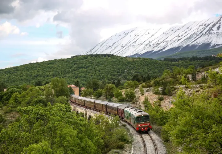 Treno storico attraversa un ponte in pietra, circondato da verde lussureggiante e montagne innevate. Un viaggio suggestivo sulla Transiberiana d'Italia.