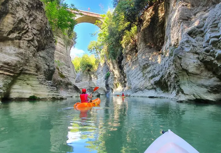 Persone in canoa navigano un fiume tra alte pareti rocciose, un paesaggio che ricorda le marmitte dei giganti, sotto un antico ponte ad arco.