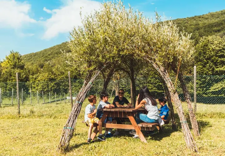 Bambini riuniti attorno a un tavolo da picnic sotto una tettoia naturale nel bosco dei folletti Urbania.