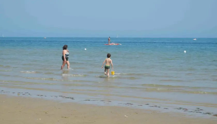 Bambino con secchiello giallo corre verso il mare a Pesaro, mentre una donna cammina nell'acqua. In lontananza, un uomo su una tavola da SUP.