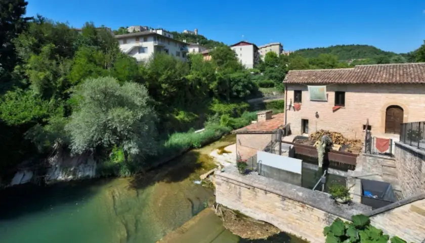 Sassoferrato cosa vedere: un antico mulino ad acqua lungo un fiume, con case e vegetazione lussureggiante sullo sfondo.