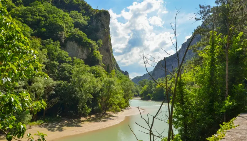 La **Gola del Furlo** vista dal fiume, con pareti rocciose ricoperte di vegetazione e un cielo azzurro con nuvole bianche.