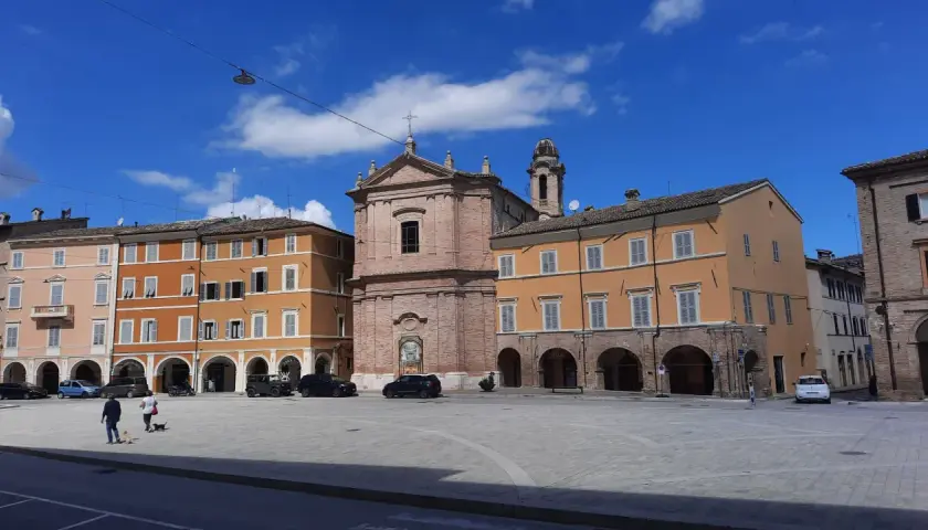 Piazza a San Severino Marche con edifici colorati, una chiesa in mattoni e cielo azzurro.