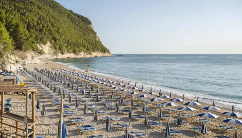 Spiaggia San Michele Sirolo: file ordinate di ombrelloni e lettini sulla sabbia dorata, con il mare azzurro e la collina verde sullo sfondo.
