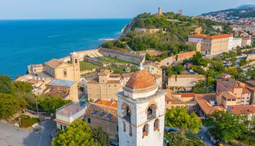 Vista aerea del **centro storico** di Ancona, con il mare blu, chiese, torri e case con tetti rossi.